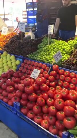 The video shows a fruit and vegetable market with various fruits and vegetables displayed in crates. A man in a white shirt and a woman in a black top are seen behind the stalls. Fruits like apples, tomatoes, bananas, peaches, apricots, and cherries are visible. Some prices are displayed on signs. The market is covered by blue and red awnings.