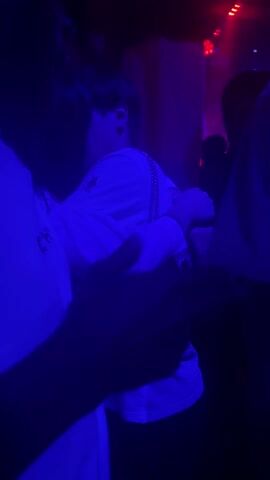 The video shows a group of people in a dimly lit environment, possibly a club. The lighting alternates between blue and red. Some individuals are wearing white t-shirts, and at least one person has a chain around their neck. The camera appears to be moving and focused on the group, but the low lighting and the people obstructing the view make it difficult to discern specific actions or interactions.
