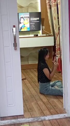 A young woman with long dark hair, wearing a black polka dot shirt and ripped jeans, sits on a wooden floor in front of a television. The television screen displays a video of two young women holding balloons. The woman on the floor is holding a white smartphone and appears to be looking at it or recording something. She has a blue clip in her hair. The room has wood-paneled walls and curtains. The scene is viewed through an open doorway with a white doorframe.