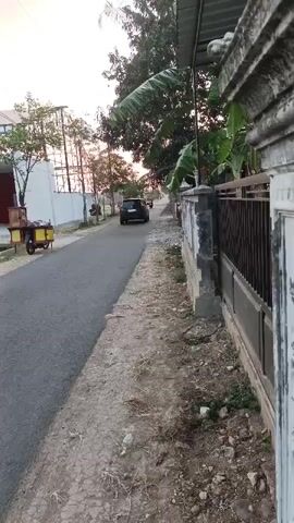 The video pans to the right, showing a residential street with a paved road and dirt shoulders. On the left side of the road, there is a building with a white facade and a corrugated metal roof, and a yellow and brown cart with wheels is parked in front of it. Further down the road, a black SUV is driving away from the camera. On the right side of the road, there is a fence with vertical wooden planks and metal sheeting, and vegetation including trees and banana leaves. The sky is light and appears to be at dusk or dawn. The overall environment suggests a rural or suburban setting.