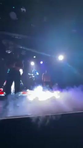 Three shirtless men are dancing on a stage lit with purple and yellow lights. The stage has a thick layer of purple fog covering the floor. The man in the center is wearing black pants, the others have their pants partially unbuttoned. A disco ball hangs from the ceiling.