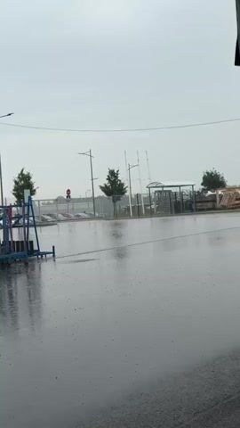 The video shows a wide-angle shot of an outdoor scene, likely a parking lot or industrial area, during a rain shower. The ground is wet and reflects the overcast sky. Several parked cars are visible in the background behind a fence. There are also trees, streetlights, and a covered structure with turnstiles. In the foreground, there is a blue metal rack with blue and clear panels. The rain can be seen as small dots on the ground and on the wet surfaces, and in the air. The lighting is dim and diffused, consistent with cloudy weather.