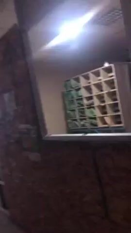 The video shows an interior space. Initially, we see a wall and a shelf with many compartments. The camera then moves through a doorway, revealing a room with fluorescent lighting. There are benches along the wall and a view out of windows. The room appears empty. The camera movement is shaky and appears to be hand-held.