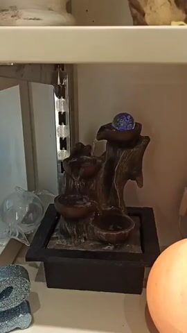 A shelf unit displays several small tabletop fountains. The first fountain, on the left, features a distressed wood-like structure with three bowls cascading water into each other, topped by a blue glass orb. To its right, another similar fountain is visible, also with cascading water. Further to the right, a larger, pink, salt lamp emits a warm glow. Below the fountains, various decorative items are visible, including a glass sphere on the far left and a pair of stacked grey stones. The video pans across the shelf, offering different views of the fountains and surrounding items.