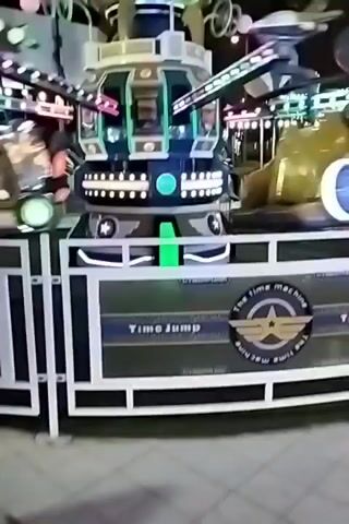 The video shows a fairground ride called "Time Jump" at night. The ride is a spinning amusement attraction with multiple arms extending from a central tower. Each arm has a pod or car at its end where riders sit. The ride is illuminated with colorful lights, including pink, blue, and green, which create streaks of light due to the motion. The central tower is also lit and appears to be decorated with a clock-like motif at the top. The ground around the ride is a paved area. In the background, other fairground attractions, including what appears to be a Ferris wheel and other rides, are visible, along with blurred figures of people. The overall impression is of a lively and exciting atmosphere at an amusement park.
