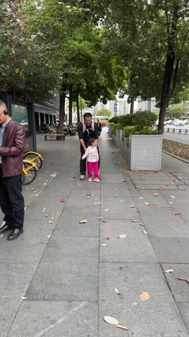 A man and a young girl are walking down a sidewalk. The man is wearing a black shirt, black pants, and a backpack. The girl is wearing a white shirt with pink sleeves, pink pants, and pink shoes. The man is smiling and holding the girl's hand. The girl is laughing and running. There are leaves scattered on the sidewalk. There are trees, buildings, bikes, and traffic visible in the background.