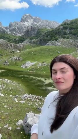 In front of a stunning mountain landscape, a woman poses in a selfie. She has long dark hair and is wearing a white hooded sweatshirt. In the background, a lake glistens, and the landscape is dotted with rocks and green fields. A few trees and what looks like a small white building are visible. The mountains rise majestically in the distance with a cloudy sky overhead. The woman gives a thumbs up to the camera.