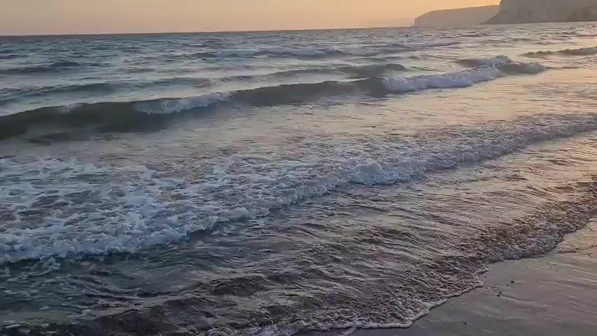 This is a drone shot following the shoreline of a beach at sunset. The water is choppy with moderate waves and foam. The sky is a hazy orange and yellow from the setting sun. In the distance, there is a cliff face. There are no people clearly visible in the frame.