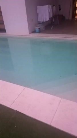 The video captures a slow pan across an empty swimming pool. The water is a clear, pale blue, with some slight ripples disturbing its surface. A light-colored tiled edge lines the pool, transitioning to a patch of green turf in the foreground. Beyond the pool, a white wall forms the background, with a wooden slatted feature visible on the left side and a glimpse of laundry hanging on a rack to the right. A small blue bucket sits near the wall. The lighting suggests it might be dusk or early morning, with a soft, diffused light. There is no visible text in the frame. The scene is tranquil and appears to be set in a residential or hotel courtyard, given the presence of the pool and the architectural elements. The overall impression is one of quiet stillness, with the empty pool waiting to be used.