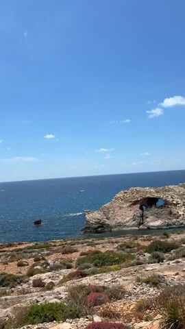 The video shows a rocky, dry coastal landscape on a sunny day with a clear blue sky dotted with small clouds. The camera pans across the scene, revealing a deep blue ocean with several boats and a distinctive rock formation with a natural arch. In the distance, a small building resembling a fort or tower sits atop a cliff overlooking the water. The foreground is covered with low-lying vegetation and rocky terrain.