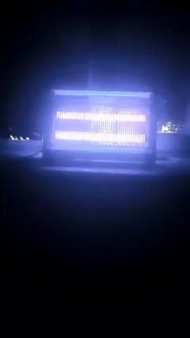 This is a video of an outdoor electric heater in the dark. The heater has multiple glowing heating elements that emit a bright orange-yellow light. The elements are arranged in horizontal rows behind a protective grid. A purple-blue light emanates from the heater's casing and the surrounding air, likely from the blue light emitted by some heating elements at certain temperatures or from residual heat. The background is dark, with some blurry lights suggesting distant buildings or streetlights. The heater appears to be positioned on a flat surface. The camera is static, capturing the heater from a medium distance.