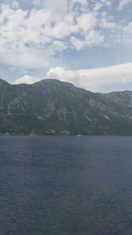 The video presents a panoramic view of a large body of water, likely a bay or lake, bordered by majestic, steep mountains. The water surface is relatively calm, showing gentle ripples across its dark blue-grey expanse. On the opposite side of the water, the imposing mountains rise sharply, covered in lush green vegetation and dotted with what appear to be small, distant settlements or houses nestled along the shoreline and lower slopes.

The sky above is partially cloudy, featuring a mix of bright blue patches and various white clouds, ranging from wispy streaks to larger, fluffy formations. The overall impression is one of natural grandeur and tranquility. The camera appears to be moving slowly, likely from a boat, offering a continuous, sweeping view of the landscape. The scene is illuminated by daylight, suggesting a time between morning and late afternoon. There are no people visible, and the focus is entirely on the natural environment. The location is Kostanjica, Montenegro.