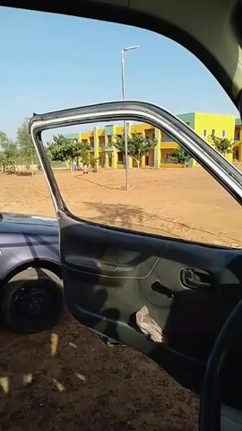 The video, filmed from inside a vehicle, shows the interior of a car door, which is open. Through the open door, a person is visible in the distance, walking in a sandy area. Beyond the person and the sandy area is a building with yellow and green sections, possibly a school or dormitory. The sky is clear and blue, indicating daytime. Another vehicle, a dark-colored car, is parked to the left of the camera, with its front tire visible. The ground in the foreground is covered in dirt and pebbles.