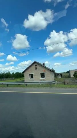 This media file captures a scenic view from a vehicle traveling along a rural road in Luzha, Russia. The scene is dominated by a vast, bright blue sky filled with fluffy white cumulus clouds. Below the sky, a lush green landscape stretches out, featuring a line of trees and a field. A metal guardrail separates the road from the field, and a wooden fence is visible in the distance. A simple wooden building with a dark roof can be seen on the right side of the frame, suggesting a rural dwelling or outbuilding. The road itself is asphalt, with distinct yellow and white lane markings. The overall impression is one of a clear, pleasant day, likely late morning or early afternoon, with good visibility and fair weather. No people are visible in the frame. The media file is a video with a duration of 5 seconds, showing the continuous movement of the landscape as the vehicle travels.