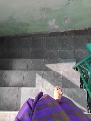 A first-person perspective video shows someone walking down a set of outdoor stairs. The person is wearing a long, purple skirt with a checkered pattern. The stairs are made of grey tiles and have green metal railings on either side. The walls are painted green, but the paint is peeling in some areas. The ground at the bottom of the stairs is also tiled.