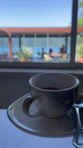 A hand places a coffee cup and saucer with a spoon on a white tablecloth. The cup contains dark coffee. Through a window behind the cup, people are sitting at tables on an outdoor patio. A lake is visible in the distance.