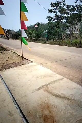 A paved road runs through a rural area. The road is lined on one side by a concrete drainage ditch, and on the other by a dirt shoulder. Along the dirt shoulder, a flagpole displays a string of triangular flags in green, yellow, red, white, and orange. Beyond the road, there are houses, trees, and vegetation. Two motorcyclists are riding on the road, one in each direction. The video is shaky and appears to be taken from a moving vehicle or by someone walking.