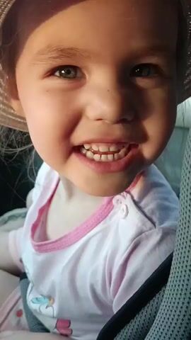 A young girl is shown in a close-up shot, seated in what appears to be a car seat. She is wearing a light-colored, round-brimmed hat with a drawstring and a light pink shirt with a white cartoon pattern and pink trim around the neck. The girl alternates between smiling, showing her teeth, and looking off to the side. The background is dark, with a textured surface next to the girl, suggesting the car seat or interior. The lighting is bright, possibly from sunlight entering the vehicle.