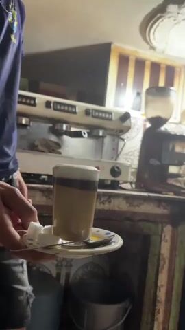 A close-up video shows a layered coffee drink being held on a saucer with two sugar cubes and a spoon. The coffee appears to be a latte or macchiato with milk foam on top. In the background, a vintage espresso machine is visible. The lighting is dim, suggesting an indoor setting, possibly a cafe or a home kitchen. The video is short, with the camera seemingly held steady as the coffee is presented.