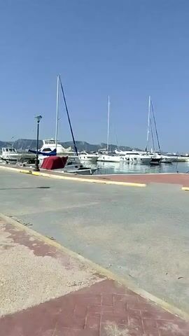 The video shows a view from a paved walkway alongside a harbor. The walkway is bordered by a concrete barrier on the left, with the ocean on the other side. In the harbor, several boats, including sailboats and a larger white boat with a red cover, are docked. Lampposts line the harbor edge. In the distance, a hilly coastline is visible under a clear blue sky. A person can be seen walking on the walkway in the distance. The camera pans slightly, showing the boats and the water.