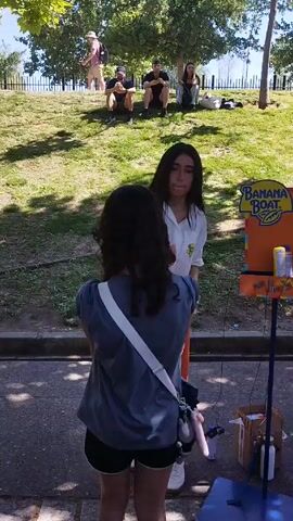 A young woman with long, dark hair is standing in front of a "Banana Boat" sunscreen display, talking to another young woman. The woman in front is wearing a white collared shirt with an orange skirt, and the woman facing her is wearing a short-sleeved blue shirt and black shorts, with a white bag strapped across her chest. In the background, people are sitting on a grassy hill. The setting appears to be an outdoor event or park.