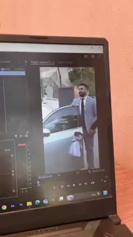 The video shows a laptop screen displaying video editing software, likely Adobe Premiere Pro. The program shows a frame of a man in a grey suit standing next to a car decorated with a white ribbon. The man is adjusting his tie and looking towards the camera. The background shows a street with parked cars. The screen shows the timeline and audio levels of the video project. The environment appears to be indoors, with a wooden surface below the laptop and a light pink wall in the background.