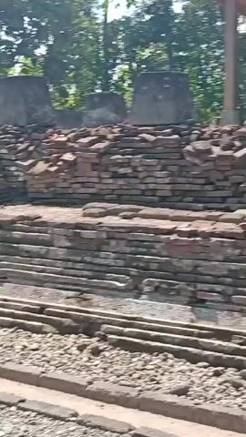 The video shows an outdoor archaeological site. In the foreground, there are piles of stones and gravel paths with concrete borders. In the background, there are brick structures and walls, some of which appear to be ruins. A small tree grows in the middle of the site. Further in the background, there are more trees and vegetation. A covered structure shelters a large, partially obscured stone statue, possibly of a Buddha, which sits on a raised stone platform. The environment appears to be warm and sunny.
