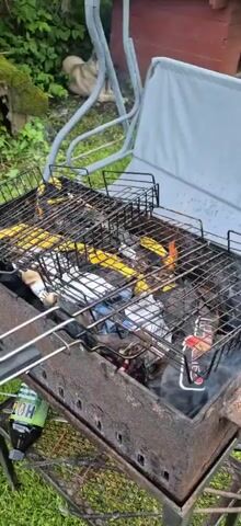 A charcoal grill is in use outdoors, with a fire burning beneath several grill baskets. The baskets contain food items, appearing to be corn on the cob and possibly some type of meat. Smoke is rising from the grill, indicating that cooking is in progress. Two bottles, one green and labeled "HOT", and another dark bottle with a white label, are visible on the ground next to the grill. The environment appears to be a grassy area with some foliage in the background, suggesting a backyard or park setting. A grey fabric cover, possibly for outdoor furniture, is partially visible in the upper right corner of the frame. The grill itself is made of dark, weathered metal and sits on a stand.