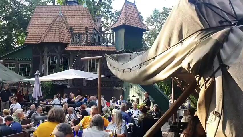 This video captures an outdoor gathering at a restaurant or beer garden with a distinct, whimsical building in the background that resembles a fairy-tale cottage or a haunted house, complete with turrets and a steeply pitched roof. The building is surrounded by lush green trees, suggesting a park-like setting. In the foreground, numerous people are seated at tables, appearing to be dining and socializing. Some tables are set with drinks and food. Large umbrellas and awnings provide shade for the patrons. The overall atmosphere seems relaxed and communal, with a large crowd enjoying the outdoor ambiance. A prominent feature in the right side of the frame is a large, draped canopy, possibly part of a tent or a covered seating area. The video pans slightly, offering a wider view of the assembled guests and the unique architecture.