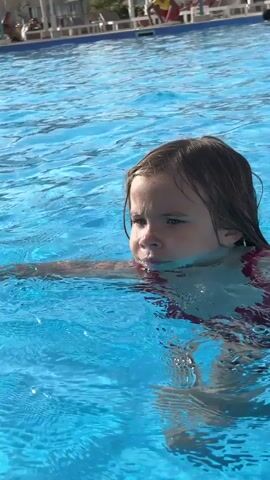 A young girl wearing a red swimsuit is in a swimming pool. She's paddling through the clear, blue water and briefly smiles. The pool is outdoors, with resort-style buildings, umbrellas, and people relaxing in the background.