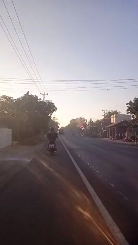 The video shows a road in a rural area at sunrise. The sun is low in the sky, casting long shadows and creating lens flares on the camera. There are several motorcycles on the road, some of which appear to be passing the camera. Trees and buildings line the sides of the road. Electrical wires are visible overhead. The overall impression is of a peaceful morning drive.