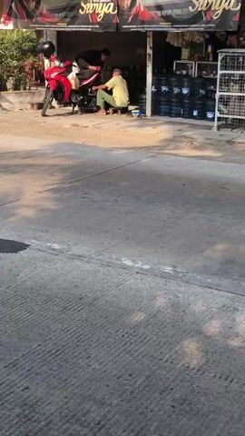 A person rides a green motorcycle with a load of yellow items strapped to the back. A white van is parked to the left. The motorcycle passes a storefront with a red sign and a person standing in the doorway. Another person on a red motorcycle is seen in the background. The scene appears to be a street in a populated area.
