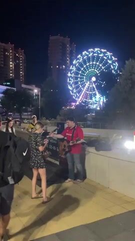 A group of people are gathered outdoors at night, possibly in an entertainment district, with a brightly lit Ferris wheel in the background. A band is performing, featuring a person playing an accordion, a woman singing into a microphone, another person playing an acoustic guitar, and a fourth person also singing. They are set up on a paved area, likely a walkway, with a low wall in the foreground. Several onlookers stand to the left of the performers, watching them. The environment suggests a public space with urban elements like tall buildings.