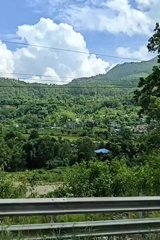 The video shows a landscape view from a moving vehicle, likely a car or bus. The foreground has a metal railing. Beyond the railing, lush green trees and vegetation dominate the scene. In the middle ground, a river flows, and scattered houses are visible among the trees on a hillside. The background features rolling green mountains under a blue sky with fluffy white clouds. Power lines are strung across the sky, and telephone poles covered in vines are also visible. The overall impression is of a scenic, natural environment with some signs of human habitation.