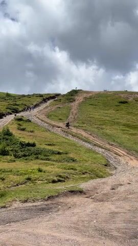 The video shows a group of people walking and biking up a steep, winding dirt path in a grassy, mountainous terrain. The path is uneven and rocky in some sections. Several individuals are seen walking in a line up the main path, while one person on a bicycle is riding down a separate, parallel path. The sky above is partly cloudy with dramatic, dark clouds. The landscape is covered in green grass and some scattered bushes and small trees. The perspective is from a slightly lower vantage point, looking up the slope.