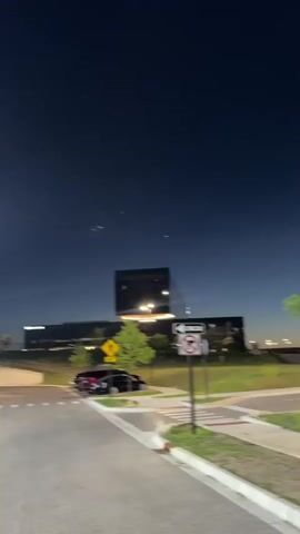 The video captures a night scene in a developed area, possibly a parking lot or street intersection, with artificial lighting from streetlights illuminating the surroundings. The sky is dark blue, indicating evening or early night.

The camera pans from left to right, revealing various elements:
- In the foreground, there are paved roads with visible crosswalk markings, curbs, and grassy areas with small trees.
- Several cars are parked along the side of the road or in parking spaces, including a prominent blue vehicle and a black SUV.
- Buildings, including a large dark structure and a long white building, are visible in the background.
- Streetlights are on, creating bright spots and some glare in the frame.
- People are visible walking through the area. At one point, two individuals are seen in a close embrace, possibly a greeting or farewell.
- Several traffic signs are visible: a yellow diamond-shaped sign with a black arrow pointing diagonally up and left, and a white square sign with a red circle and a diagonal line across it, featuring a black arrow pointing to the right.

The video has noticeable motion blur, suggesting the camera is moving or the subjects are in motion, contributing to a dynamic yet somewhat indistinct visual quality. The overall impression is that of a bustling area at night, with both vehicles and pedestrians present.