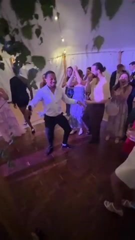 A man in a white shirt and black pants dances and sings into a microphone on a wooden dance floor. He is surrounded by people in formal attire dancing. The event appears to be a wedding reception, with string lights and greenery overhead. Some guests are holding drinks and appear to be enjoying themselves. The camera is shaky, suggesting it is handheld.