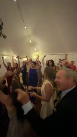 A wedding reception is underway inside a tent, with string lights overhead and greenery adorning the tent poles. Many people are dancing, with some raising their arms in the air. In the center of the frame, one man is being carried on the shoulders of another man while dancing. Several individuals are holding up their phones to record the event.