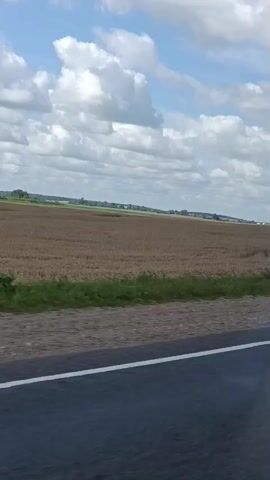The video shows a view from a moving vehicle, looking out over a large, harvested agricultural field. The field is mostly brown and appears to have stubble from a recently harvested crop. A strip of green grass runs along the edge of the field closest to the road. The road itself is visible in the foreground, with a white line marking the edge. The sky is partly cloudy with scattered white clouds against a blue sky. In the distance, there are some trees and buildings, suggesting a rural or semi-rural area. The lighting indicates it is daytime. There are no people or animals visible in the frame.