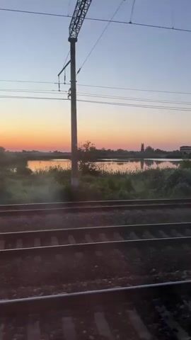 This video shows a view from a moving train at sunset. The camera is positioned to capture the train tracks in the foreground, running horizontally across the frame. Beyond the tracks, there is lush green vegetation and a body of water. The sky is a gradient of blue and orange, indicating the setting sun. Overhead, power lines and a utility pole are visible, typical of a railway environment. The scene is peaceful and depicts a journey through a natural landscape during golden hour.