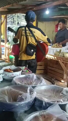 A man in a yellow shirt with a black bag slung across his shoulder is playing an acoustic guitar. He is standing in front of a market stall with various food items displayed. Another man is working at the stall behind him, preparing produce. The environment appears to be an outdoor market with a corrugated metal roof. The video captures a moment of live music performance in a commercial setting.