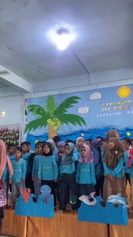 A group of children, most wearing blue shirts and headscarves, stand on a stage in front of a blue banner decorated with a palm tree, clouds, and the words "PERPISAHAN IES SCHOOL ANGKATAN KE 8". Several of the children are holding microphones and appear to be singing or speaking. Some children have their hands clasped in front of them, while others gesture with their hands. A few adults, also wearing headscarves, stand behind the children, with one adult to the right of the frame speaking into a microphone. In front of the children, there is a stage decorated with blue cutouts resembling ocean waves and sea creatures, including a jellyfish, an octopus, a fish, and dolphins. The lighting in the room is bright, with a ceiling light visible at the top of the frame. The overall scene depicts a school event or performance.