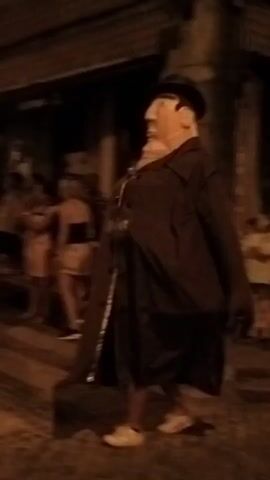 A group of people, some dressed in what appear to be matching outfits with circles on them, are gathered on a cobblestone street at night. Some of the individuals are holding and playing drums. One person in the foreground is wearing a dark robe with a hood. Streetlights illuminate the scene. There are buildings lining the street. The mood appears to be festive.