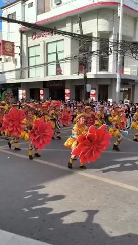 A color video shows a large group of dancers performing in the street, during a parade. They all wear colorful patterned outfits in yellow, orange and black, and matching headdresses topped by large red flowers. They walk toward the camera in rows, performing dance moves. A crowd of spectators watches from the sidewalk. Buildings are visible on both sides of the street, and streetlights and power lines hang overhead. Signs for McDonald’s and Chowking are visible.