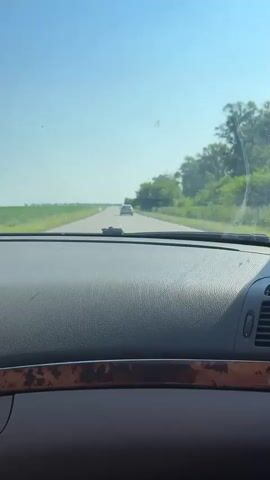 The video begins with a view from inside a vehicle looking out the windshield. The dashboard is visible in the foreground. A light-colored car is driving on a paved road ahead, and it appears to be slightly off-center to the left. The road stretches into the distance, with grassy fields on either side. Trees and foliage line the right side of the road. The sky is clear and blue. The car in front is moving away from the camera, and as the video progresses, the camera car appears to be gaining on it.