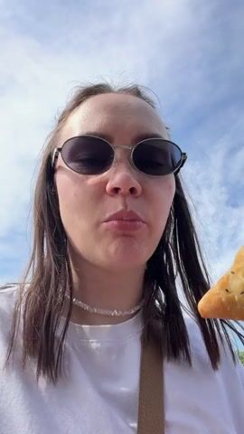 A young woman with dark hair and sunglasses is featured in the video. She wears a white t-shirt and a light-colored beaded necklace.

In the first shot, she is seen taking a bite out of a triangular baked pastry, which is topped with black seeds. The pastry is held in her right hand. The camera focuses on her mouth as she chews. The sky above her is bright blue with scattered white clouds, indicating daytime.

The video then cuts to a slightly wider shot where the woman smiles at the camera, still holding the pastry. She is standing outdoors next to the open trunk of a car. In the background, a house with a red roof and a grassy area can be seen under the clear, sunny sky, suggesting a rural or village setting. She appears happy and content. In this shot, a small bottle is visible inside the car trunk, and she wears a wristwatch on her left wrist.