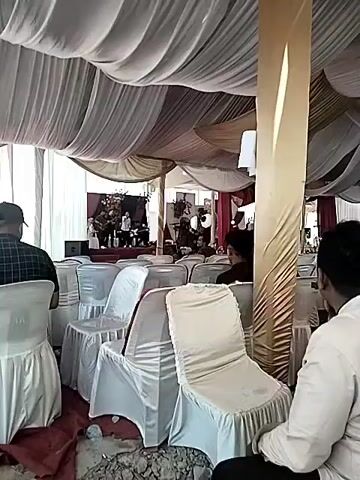The video shows a gathering under a large tent. People are seated in white chairs, some of whom are looking toward a stage area. The tent ceiling is draped with white and gold fabric. A man in a plaid shirt holds up a small cup with a design on it and smiles at the camera.