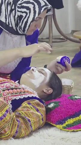 A woman in a blue tunic and a black and white striped headscarf is applying white cream to the face of a younger person lying down. The younger person is wearing a heavily embroidered, colorful garment and is lying on a brightly colored woven mat. The woman holds a small, purple jar of cream in her left hand. The scene appears to be indoors, with a wooden chair visible in the background.