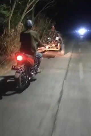 A person wearing a helmet and a green shirt is riding a red motorcycle at night. The motorcycle is following a small tractor that is pulling a trailer loaded with branches and possibly other debris. The tractor is in front of the motorcycle, and its headlights are illuminating the road ahead. The scene is dark, with trees and foliage visible on the sides of the road. The road appears to be paved and has white lane markings. The overall impression is of a rural setting at night, with the tractor and motorcycle traveling together.