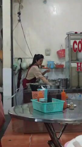 A woman in a tan shirt and red apron is working behind a counter in what appears to be a small restaurant or food stall. She uses a ladle to scoop something from a large metal pot. Behind her, various containers and utensils are visible, along with a glass display case labeled "Cơm H." A shiny metal table in the foreground holds a turquoise basket filled with condiments, along with other small containers. The environment looks simple and utilitarian, with white walls and basic fixtures.